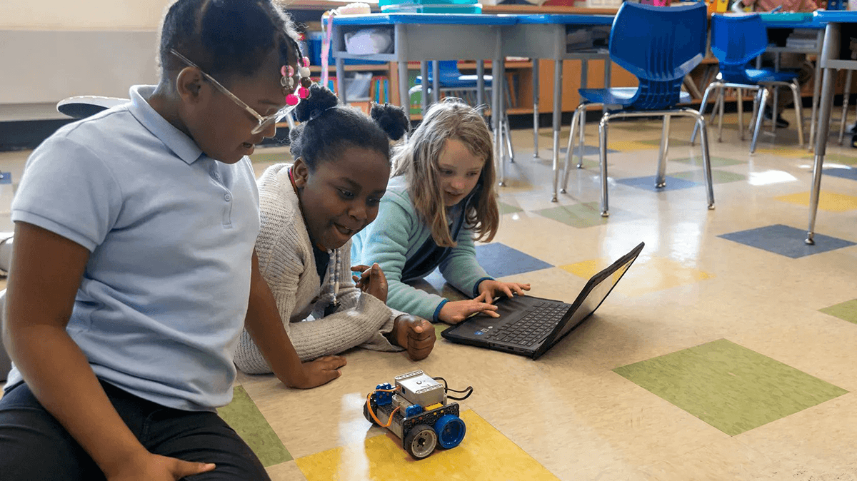 Three girls sit on the floor with a laptop and a VEX GO Code Base Robot. One has an excited expression on her face as she watches the robot move.