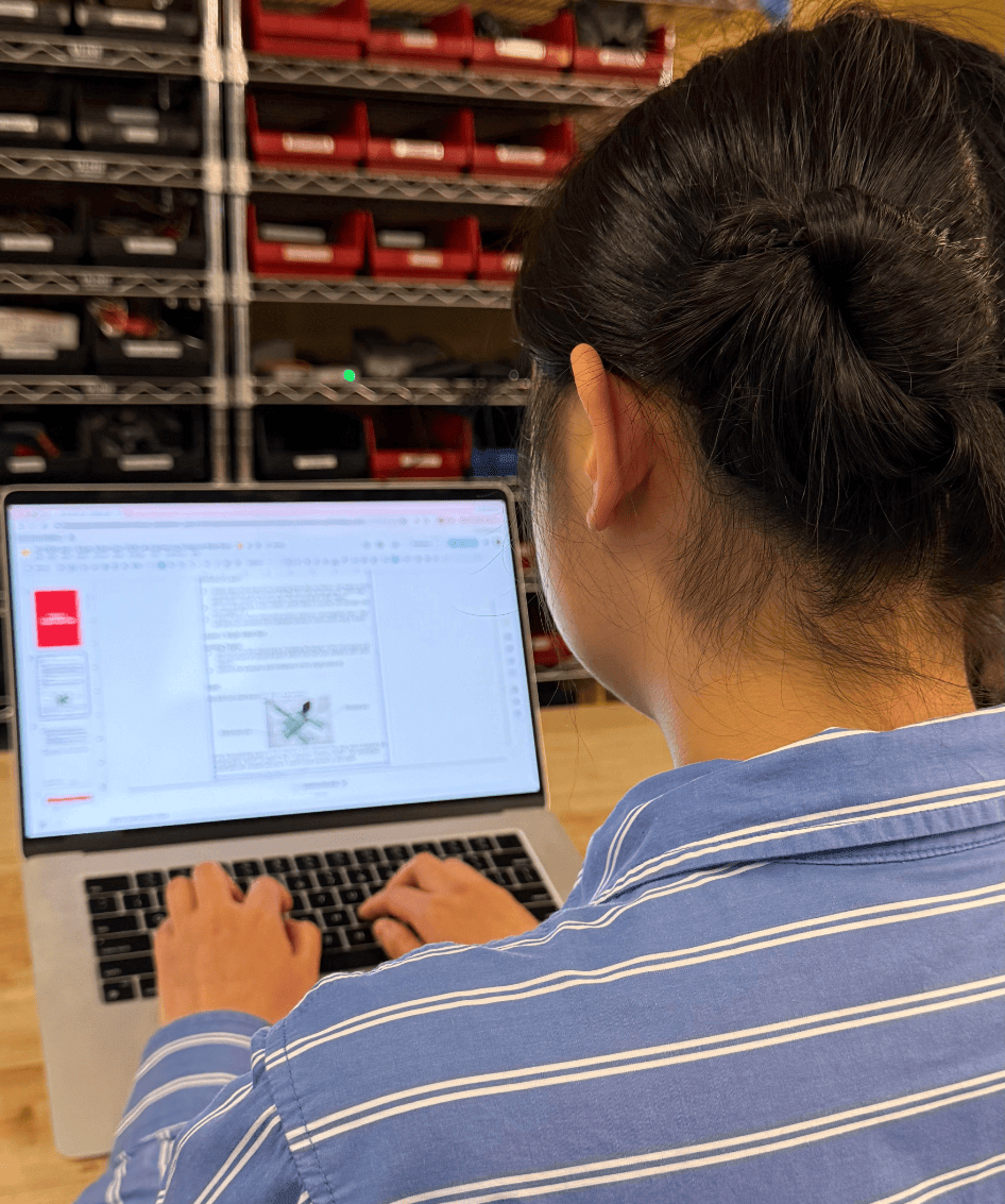 Over the shoulder view of a student working on a digital engineering notebook on a laptop. There are shelves of VEX Parts shown in the background.