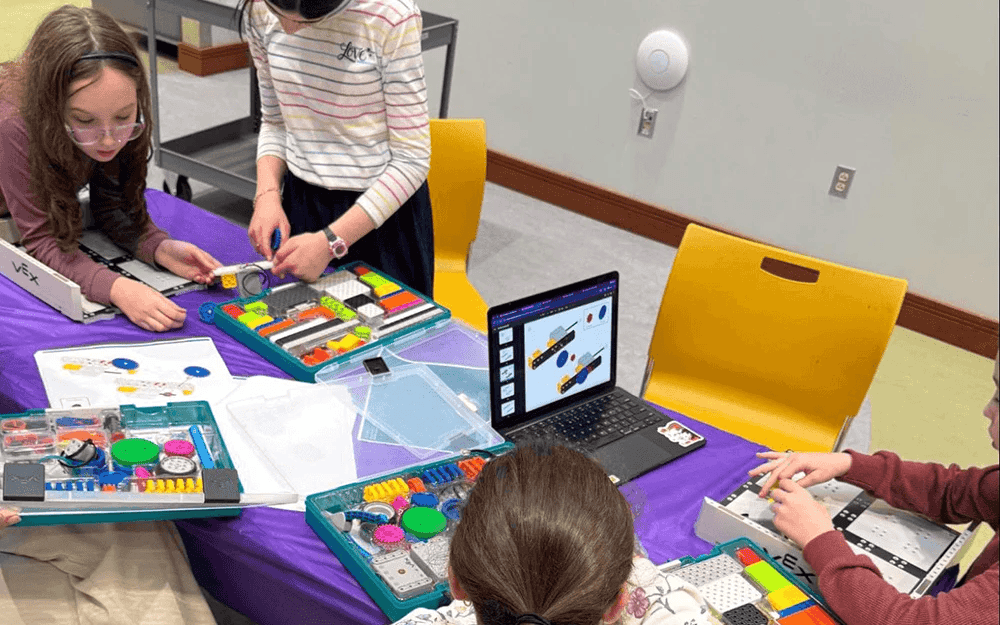 Students working together at a table with VEX GO kits, papers, and build instructions on a laptop.