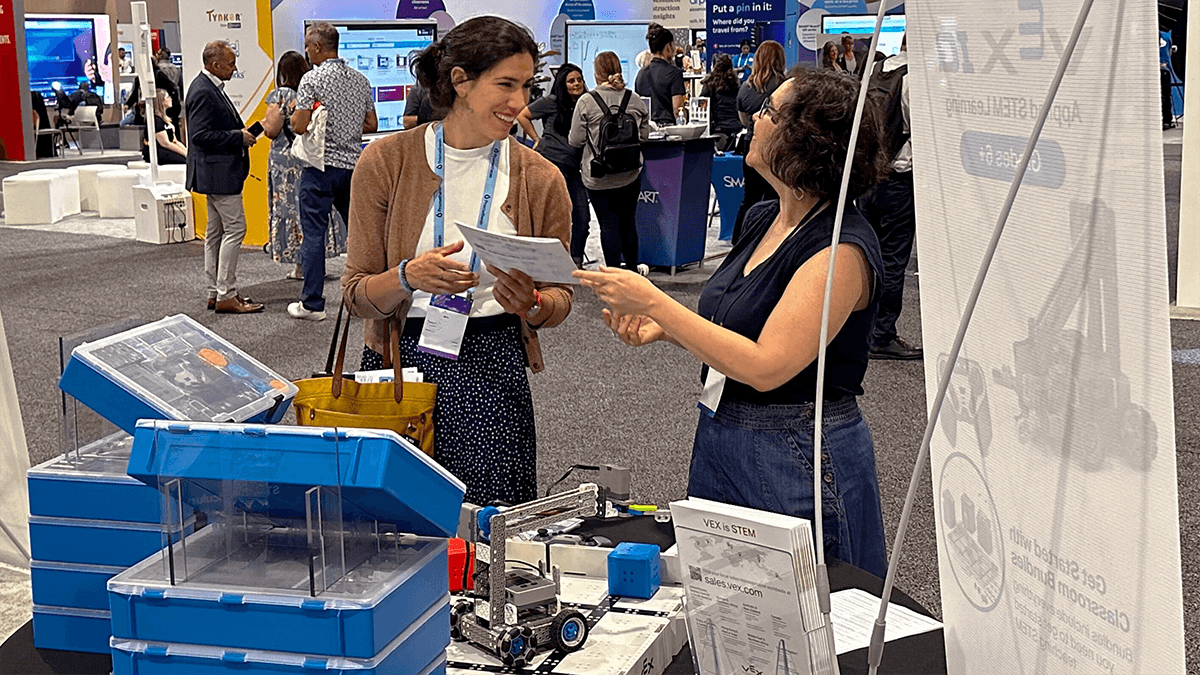 Two educators talking with each other at a conference next to a table displaying VEX IQ Kits, robots, and documents.