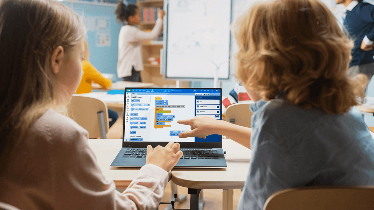 Kids in a classroom pointing at a computer with a VEXcode Blocks project open.