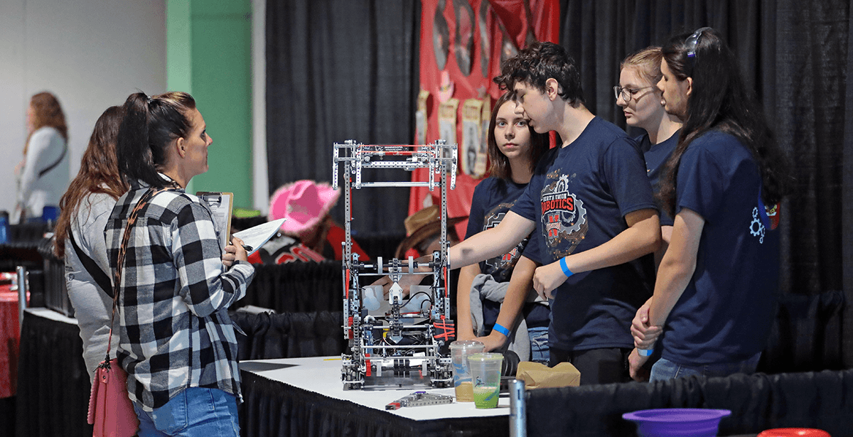 A VEX V5 team showing their robot on a table in their pit during an interview with two people.